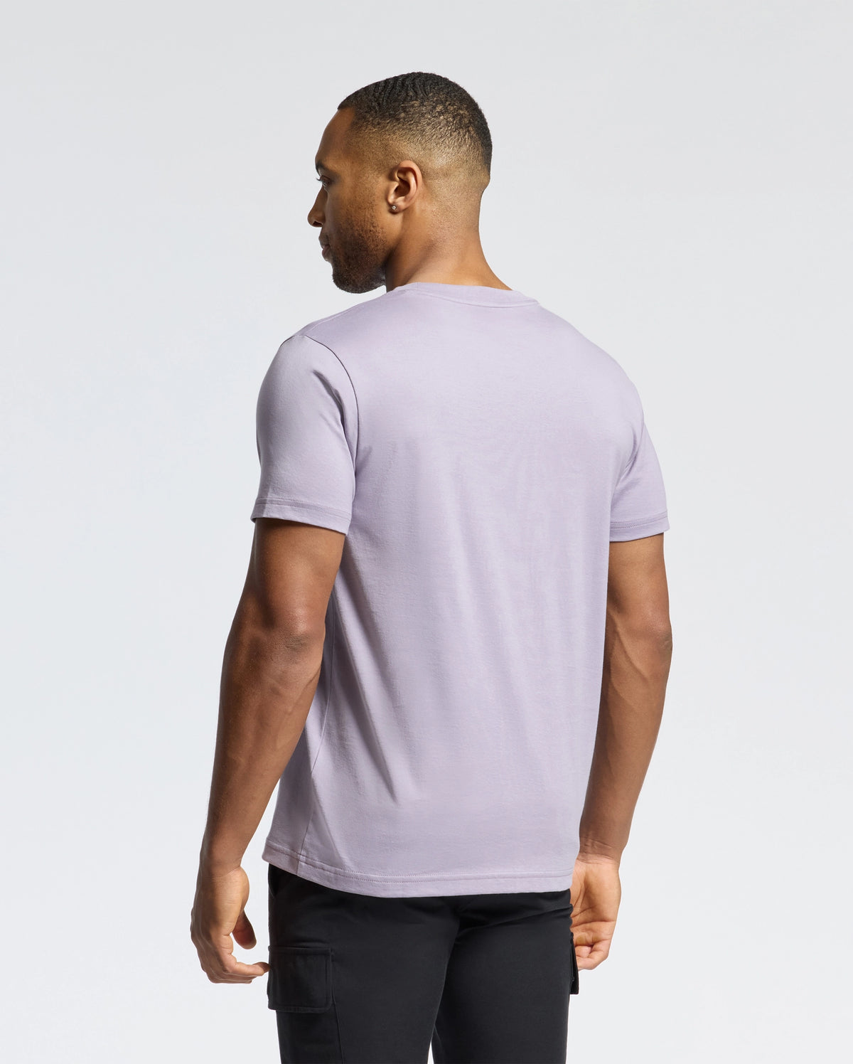 A short-haired individual wears a light purple MENS JAMES CREW NECK TEE by Psycho Bunny and black pants, facing away from the camera against a plain white backdrop.