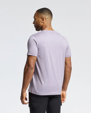 A short-haired individual wears a light purple MENS JAMES CREW NECK TEE by Psycho Bunny and black pants, facing away from the camera against a plain white backdrop.