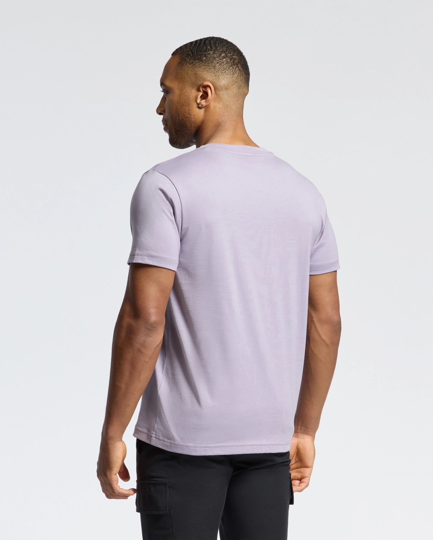 A short-haired individual wears a light purple MENS JAMES CREW NECK TEE by Psycho Bunny and black pants, facing away from the camera against a plain white backdrop.