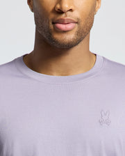 A person wears the Psycho Bunny MENS JAMES CREW NECK TEE, made of lavender Pima cotton, featuring a stylized rabbit and crossed bones logo on the chest. Only their lower face and chest are visible, highlighting its crease-resistant collar.