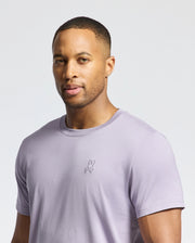 Wearing the Psycho Bunny MENS JAMES CREW NECK TEE (B6U952D200) in light purple, a person with short hair stands against a plain backdrop. The tee features an elegant embroidered logo on the chest as they gaze directly at the camera.