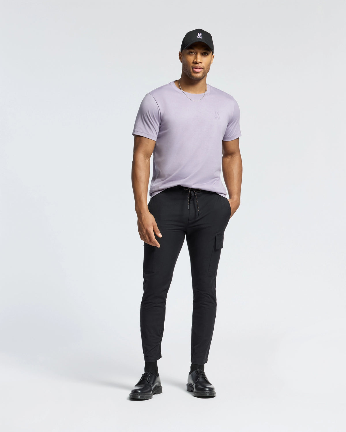 A man stands against a white background in a black cap, black jogger pants, and shoes. Wearing the Psycho Bunny MENS JAMES CREW NECK TEE in light purple Pima cotton with hands in pockets, he strikes a casual pose exuding luxe comfort.