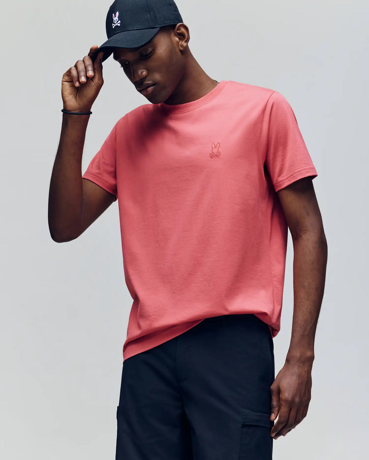 A man in a black cap and the Psycho Bunny MENS JAMES PREMIUM CREW NECK TEE (B6U952D200), featuring coral pink Pima cotton with premium embroidery, stands against a plain background, tilting his head and touching his cap's brim.
