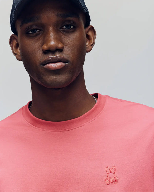 A man in a black cap and Psycho Bunny's MENS JAMES PREMIUM CREW NECK TEE, featuring a pink Pima cotton build and the signature bunny and crossbones logo, poses against a gray background, showcasing the tee's premium embroidery.