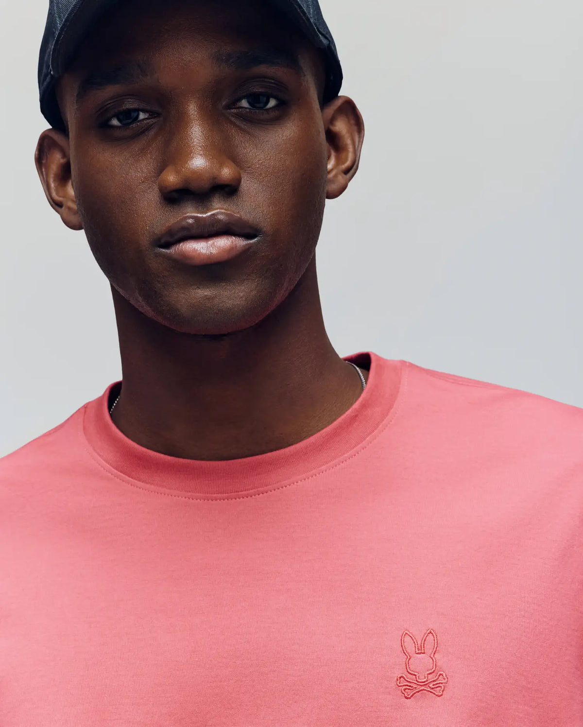 A man in a black cap and Psycho Bunny's MENS JAMES PREMIUM CREW NECK TEE, featuring a pink Pima cotton build and the signature bunny and crossbones logo, poses against a gray background, showcasing the tee's premium embroidery.