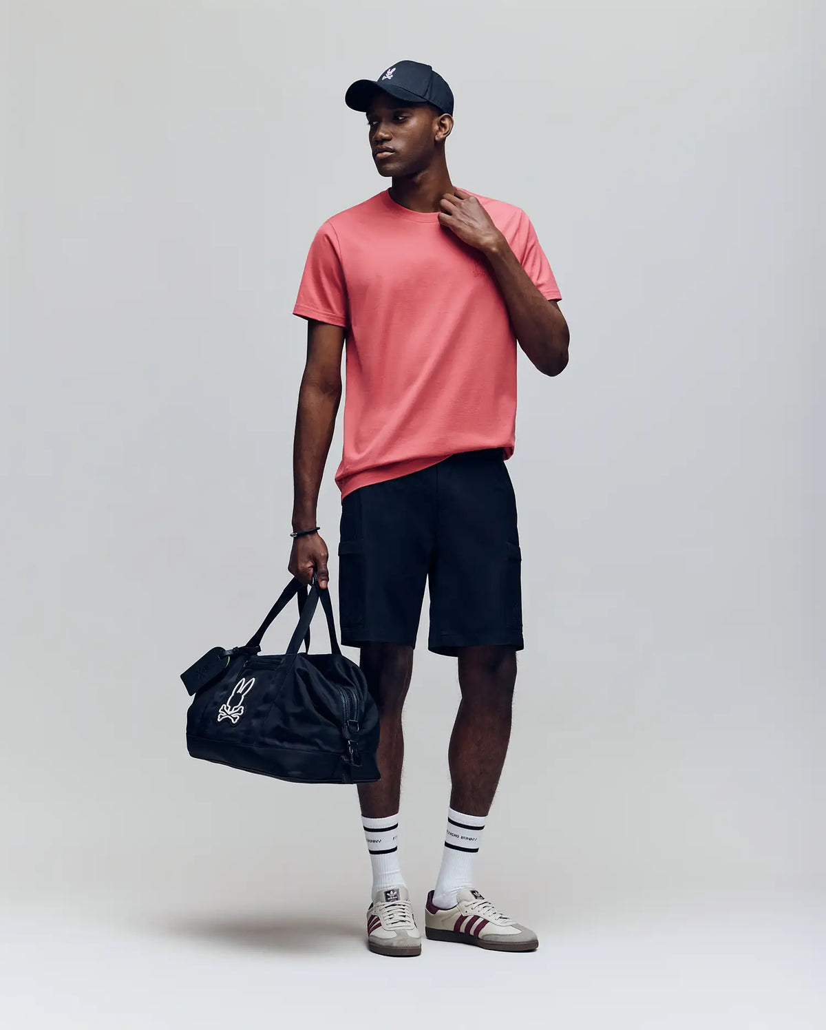 A man stands against a plain background wearing the Psycho Bunny MENS JAMES PREMIUM CREW NECK TEE in red, paired with a black cap, black shorts, striped white socks, sneakers, and holding a black duffel bag with a white logo.