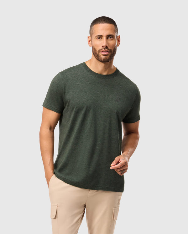 A man with short hair and a beard is wearing a dark green, stylish MENS JAMES PREMIUM CREW NECK TEE from Psycho Bunny, made of premium Pima cotton, along with beige cargo pants. He stands against a plain light gray background.