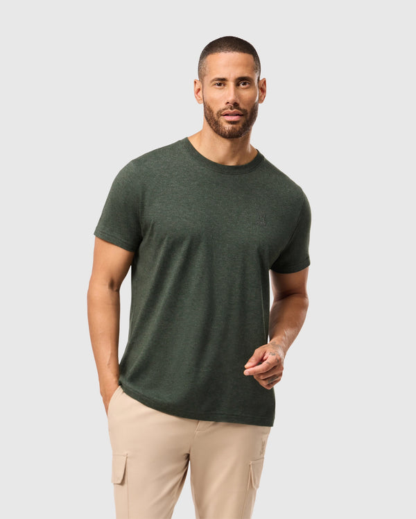 A man with short hair and a beard is wearing a dark green, stylish MENS JAMES PREMIUM CREW NECK TEE from Psycho Bunny, made of premium Pima cotton, along with beige cargo pants. He stands against a plain light gray background.
