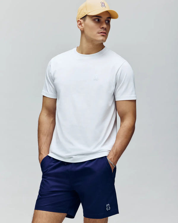 A man in a pale yellow cap, navy blue shorts, and a Psycho Bunny MENS JAMES PREMIUM CREW NECK TEE (B6U952D200) stands with hands in pockets, looking to the side against a plain grey background.