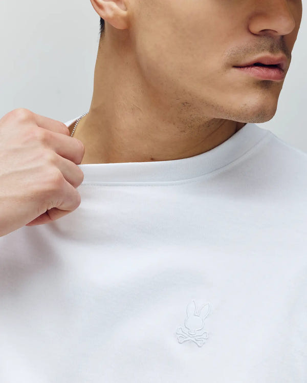 A person in the MENS JAMES PREMIUM CREW NECK TEE by Psycho Bunny, made from Pima cotton and featuring an embroidered bunny and crossbones logo on the chest, holds the collar, partially revealing their face and neck.