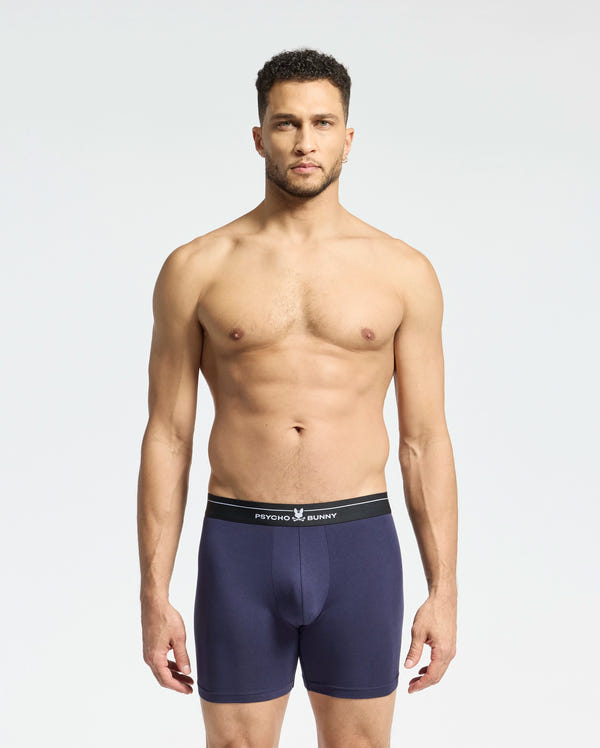 A man stands against a plain background wearing navy blue Psycho Bunny boxer briefs with a black waistband. Made from stretch Pima cotton, these breathable briefs promise supreme comfort. He looks directly at the camera with a neutral expression. Product: MENS 2 PACK BOXER BRIEF - B6V481F200.