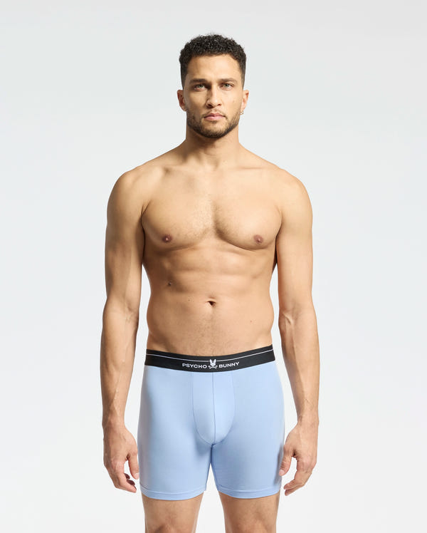 Against a plain backdrop, someone models light blue breathable boxer briefs with a black waistband. "Psycho Bunny" is on them. Crafted from stretchy Pima cotton, they offer daily comfort. The person has short hair and looks straight ahead, wearing the MENS 2 PACK BOXER BRIEF - B6V481F200.