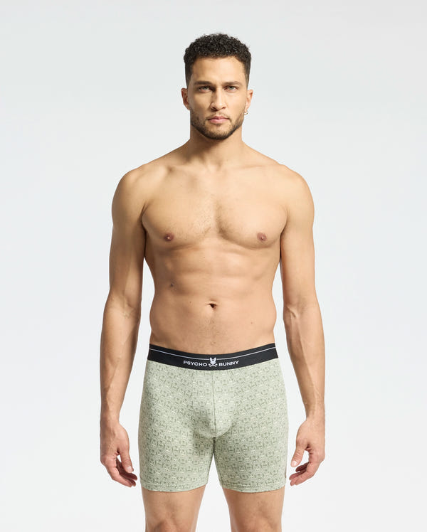 A man stands confidently in Psycho Bunny's MENS 2 PACK BOXER BRIEF - B6V481F200, crafted from a Pima cotton-Modal blend. These boxer briefs feature a subtle pattern and contrasting black waistband with the brand logo, designed for all-day comfort. He poses against a light gray background.