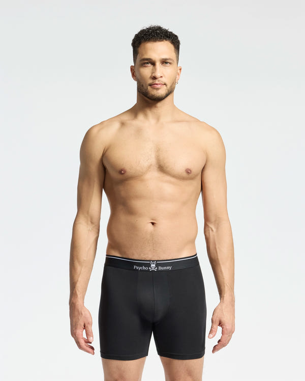 A man stands facing forward in MENS SOLID KNIT 2 PACK BOXER BRIEF - B6V906ARCM by Psycho Bunny, featuring an ultrasoft modal-blend and a branded waistband. The background is plain and light-colored.