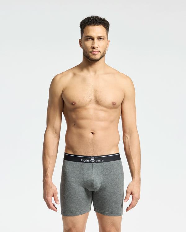 A person stands against a plain, light background wearing Psycho Bunny's gray MENS SOLID KNIT 2 PACK BOXER BRIEF - B6V906ARCM made from Pima cotton, featuring a black waistband with "Psycho Bunny" text. They look directly at the camera with a neutral expression.