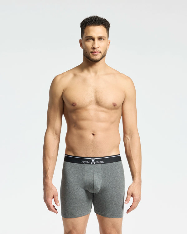 A person stands against a plain, light background wearing Psycho Bunny's gray MENS SOLID KNIT 2 PACK BOXER BRIEF - B6V906ARCM made from Pima cotton, featuring a black waistband with "Psycho Bunny" text. They look directly at the camera with a neutral expression.