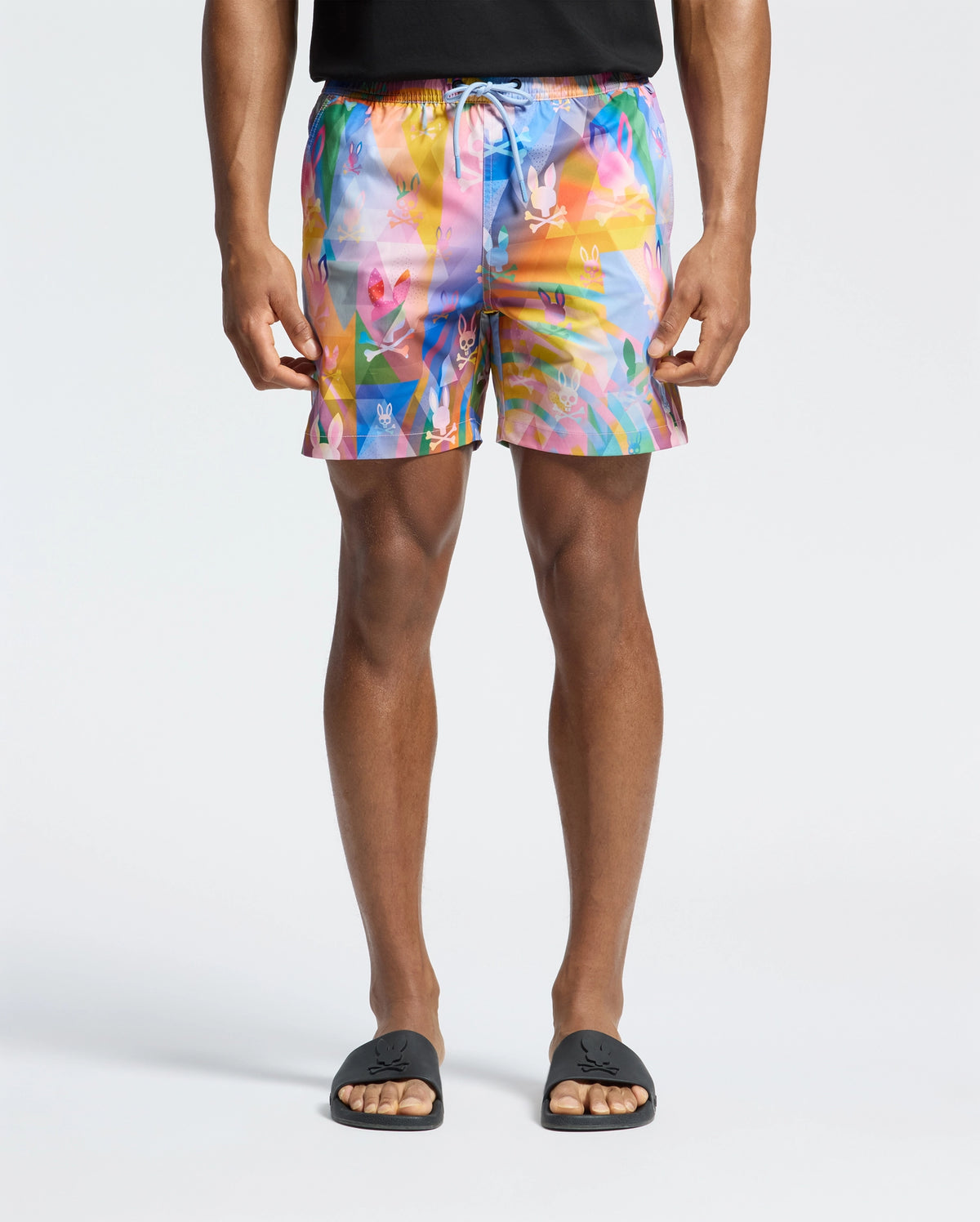 Sporting the Psycho Bunny MENS MILOS PRINTED SWIM TRUNK - B6W446F200, a person showcases vacation style with multicolored abstract-patterned shorts in pink, blue, yellow, and orange. Paired with a black shirt and sandals on a white backdrop, these quick-dry shorts offer ultimate comfort.