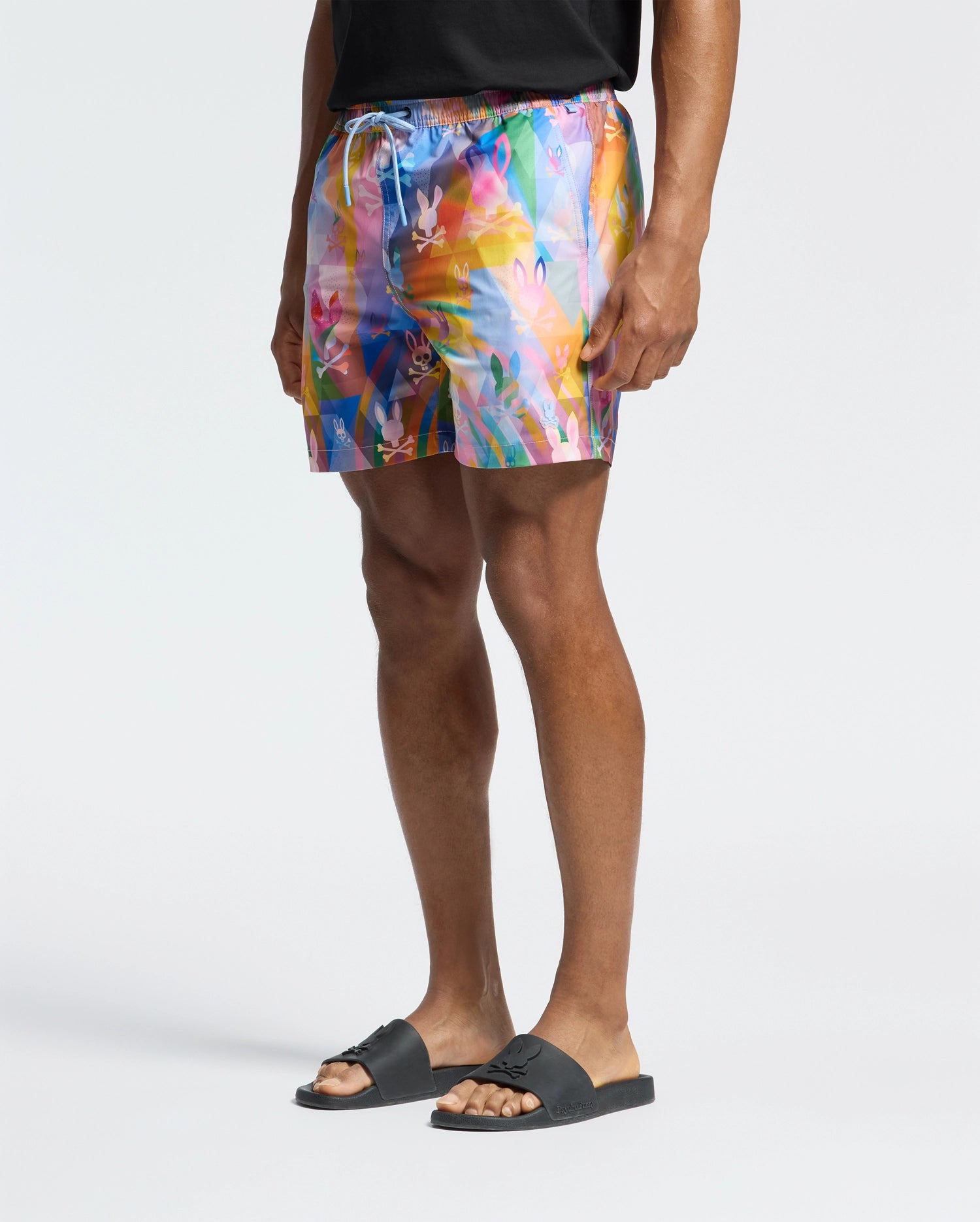 A person in a black t-shirt, Psycho Bunny's MENS MILOS PRINTED SWIM TRUNK - B6W446F200, and black sandals stands against a plain background, capturing a relaxed vacation vibe.