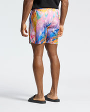Showcasing the Men's Milos Printed Swim Trunk - B6W446F200 by Psycho Bunny, a person is seen in vibrant shorts with abstract bunny designs, ideal for vacation style. Quick-dry fabric ensures comfort, complemented by a black shirt and slides against a plain background.