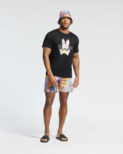 A man radiates vacation style in a black t-shirt with a vibrant print, pairing Psycho Bunny's MENS MILOS PRINTED SWIM TRUNK - B6W446F200 rainbow tie-dye shorts, a matching bucket hat, and black slide sandals against a plain white backdrop.