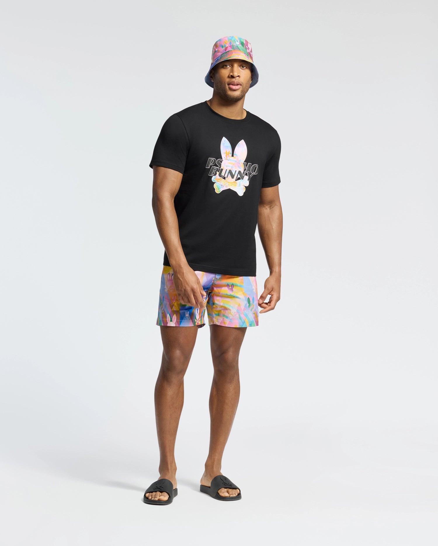A man radiates vacation style in a black t-shirt with a vibrant print, pairing Psycho Bunny's MENS MILOS PRINTED SWIM TRUNK - B6W446F200 rainbow tie-dye shorts, a matching bucket hat, and black slide sandals against a plain white backdrop.