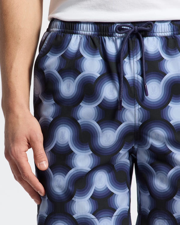 A person is modeling Psycho Bunny's Mens Paros Printed Swim Trunk, style B6W450F200, in navy blue and light gray with geo patterns and wavy circular designs. The swim shorts feature a drawstring waist and are displayed waist-to-mid-thigh with the right hand relaxed by their side.