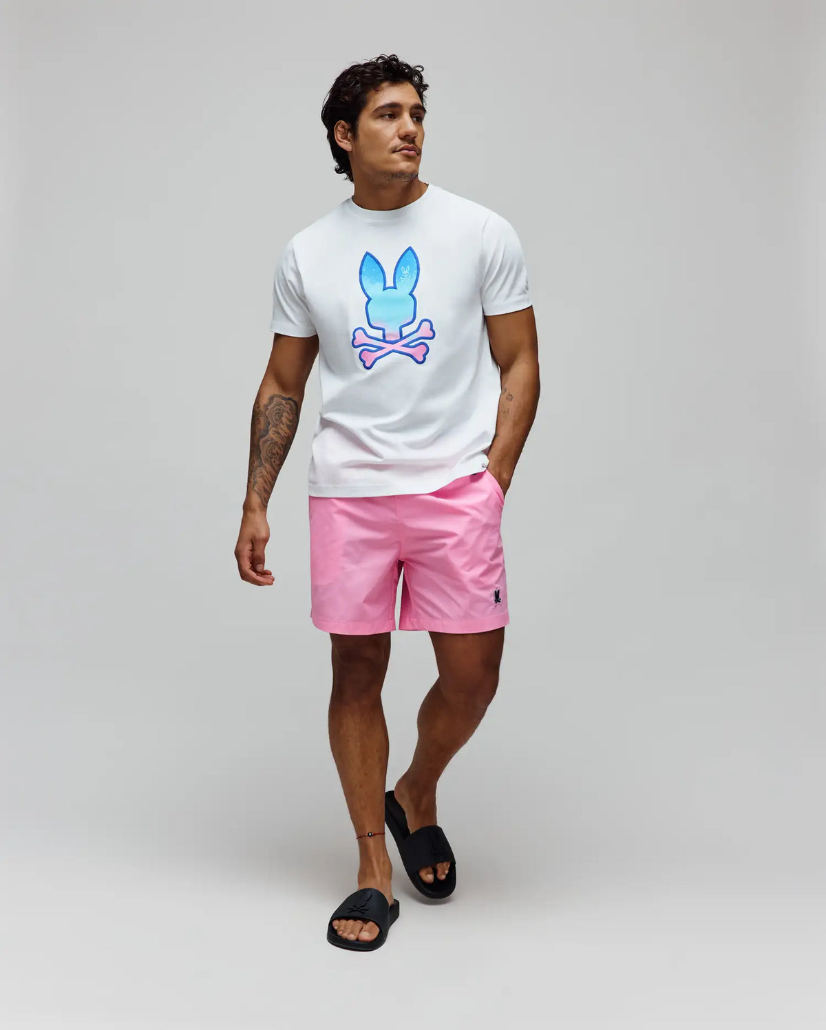 White jersey t-shirt with a large blue-to-pink gradient Psycho Bunny skull graphic on the chest, paired with Mens Samos Hydrochromic Printed Swim Trunk in pink, featuring a small dark embroidered bunny logo. (6)