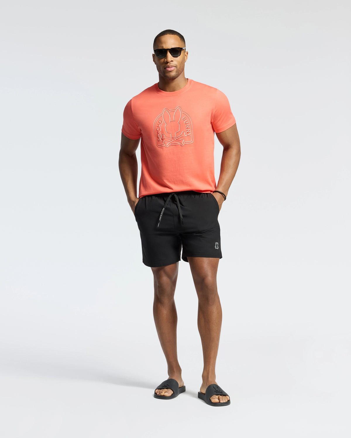 Coral jersey t-shirt with a large outlined Psycho Bunny skull logo on the chest, paired with black Mens Samos Hydrochromic Printed Swim Trunk featuring a white drawstring and small white bunny skull logo on the left leg.