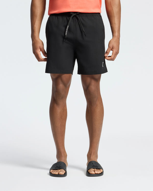 The image shows a person wearing Psycho Bunny's black Samos Hydrochromic Printed Swim Trunk with a small left-leg logo, a coral shirt, and black slide sandals, focusing on their lower body.