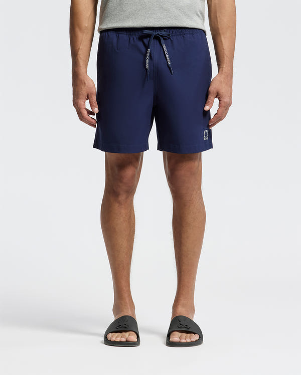 A person stands against a plain light background, wearing Psycho Bunny MENS SAMOS HYDROCHROMIC PRINTED SWIM TRUNK - B6W453F200 in navy blue; only their lower body is visible.