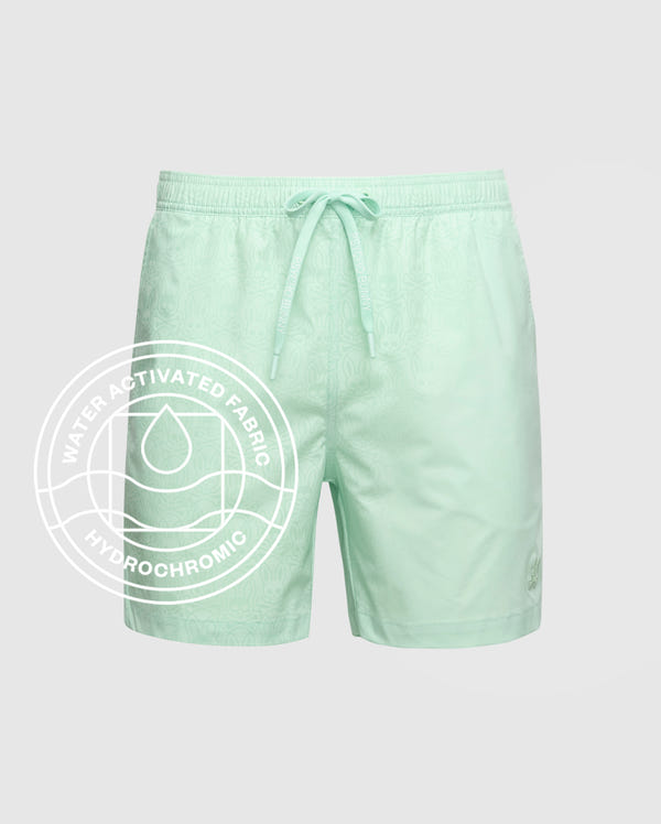 The Psycho Bunny MENS SAMOS HYDROCHROMIC PRINTED SWIM TRUNK - B6W453F200 features a light green subtle pattern, elastic waistband, and drawstring. Water-activated hydrochromic fabric reveals a white circular graphic with droplet icon when wet.