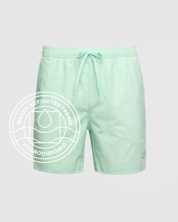 The Psycho Bunny MENS SAMOS HYDROCHROMIC PRINTED SWIM TRUNK - B6W453F200 features a light green subtle pattern, elastic waistband, and drawstring. Water-activated hydrochromic fabric reveals a white circular graphic with droplet icon when wet.