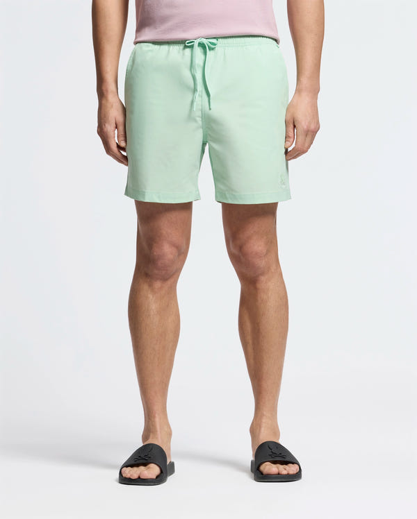 A person is dressed in Psycho Bunny's MENS SAMOS HYDROCHROMIC PRINTED SWIM TRUNK - B6W453F200 with a light green and bunny pattern, black slide sandals, and a pale pink top, standing against a plain background.