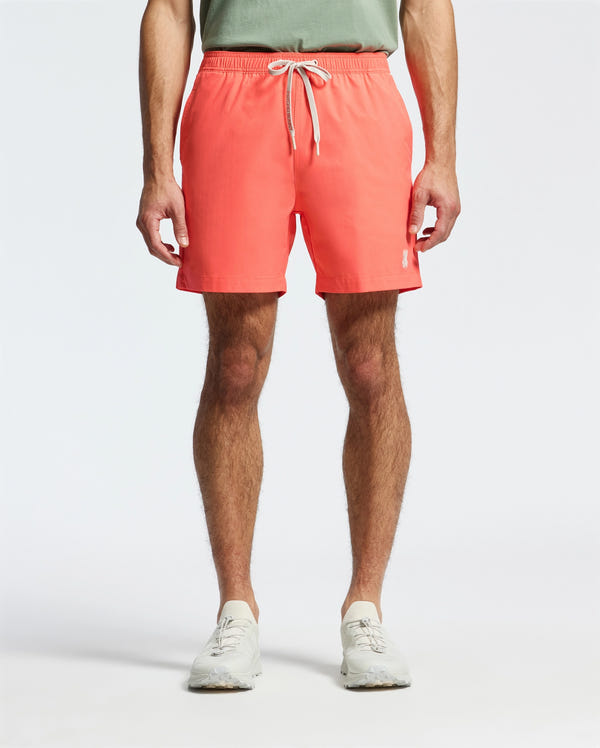 The individual poses in coral Psycho Bunny MENS SAMOS HYDROCHROMIC SWIM TRUNK with a white drawstring against a white backdrop, paired with a light green shirt and sneakers. The focus is on the trendy swim trunks, ideal for any pool party atmosphere.