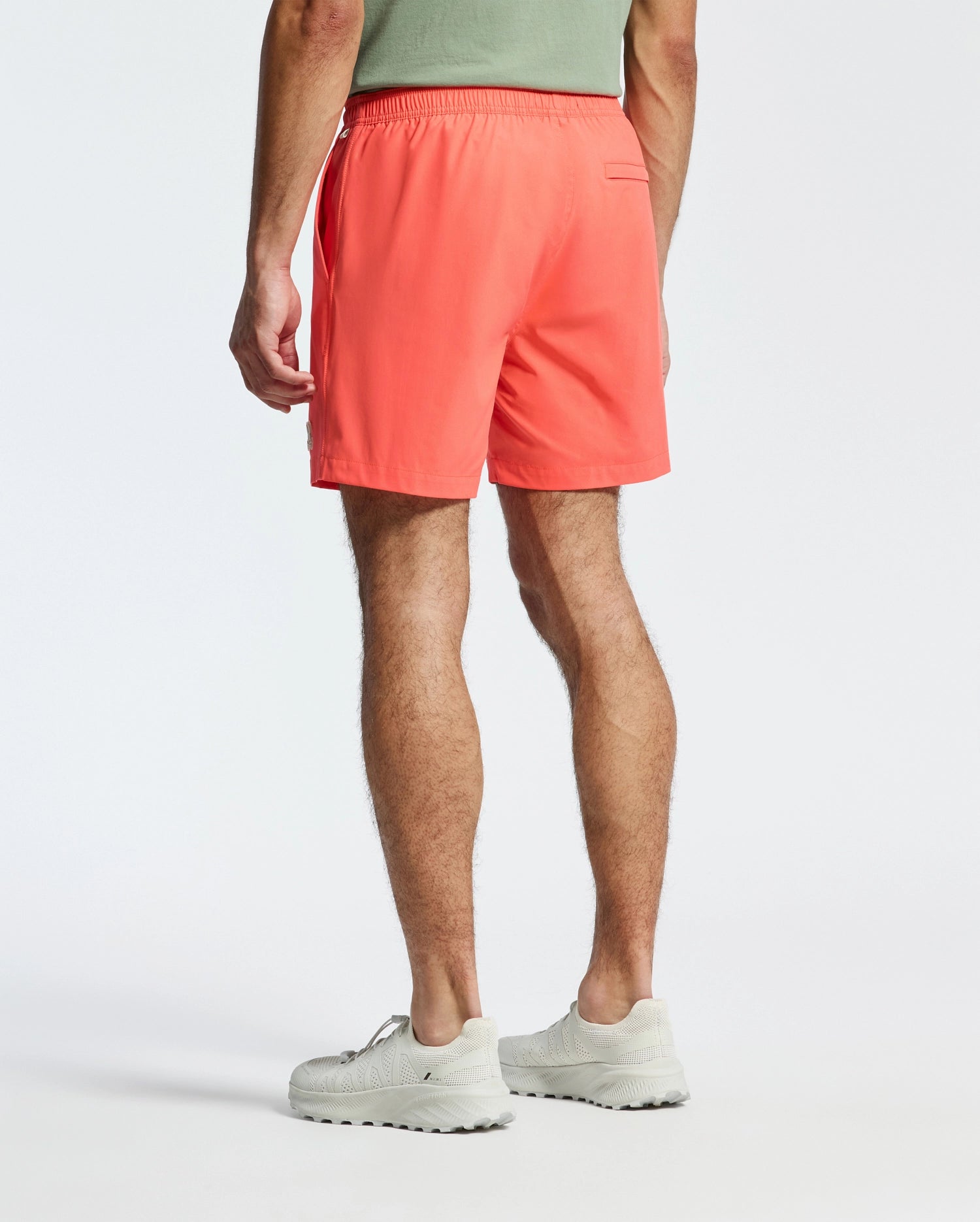 A person is wearing Psycho Bunny's MENS SAMOS HYDROCHROMIC SWIM TRUNK in coral and a green shirt, showcasing a back view. They have light-colored sneakers on, standing against a plain white background, all set for a pool party.