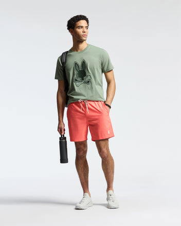 A man in a green Psycho Bunny T-shirt and MENS SAMOS HYDROCHROMIC SWIM TRUNK - B6W453F200 holds a black hydrochromic bottle, sports white sneakers, and has a striped strap over his shoulder against a light gray backdrop, ready for the pool party.