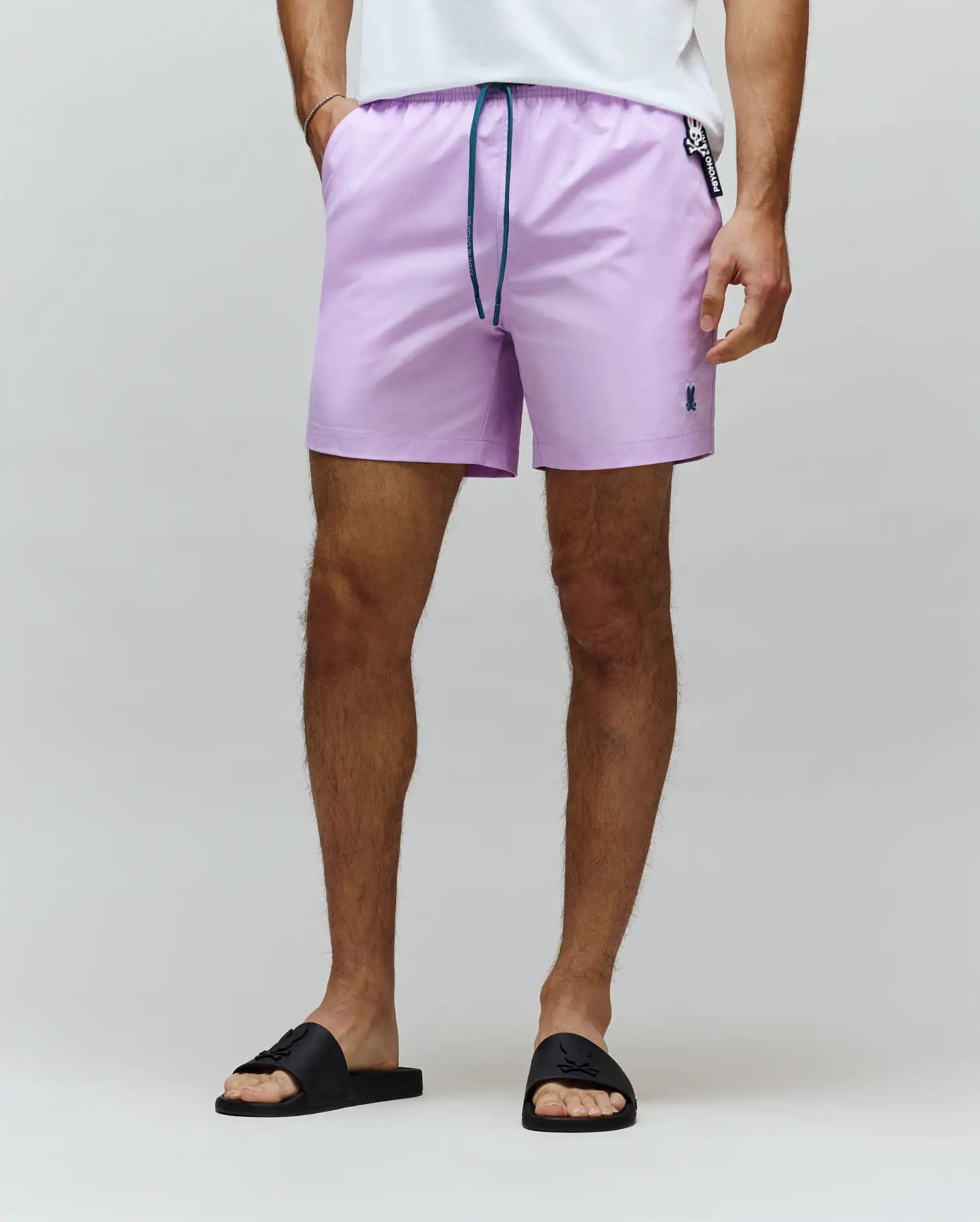 Mens Samos Hydrochromic Printed Swim Trunk, lavender with a classic fit, smooth fabric, teal drawstring, small dark blue embroidered Psycho Bunny skull logo on the left leg, and a branded tag.