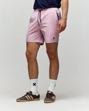 A person wearing Psycho Bunny MENS SAMOS HYDROCHROMIC PRINTED SWIM TRUNK - B6W453F200 in light pink, a navy shirt, white crew socks with a small black logo, and brown Adidas sneakers stands against a plain light gray background.