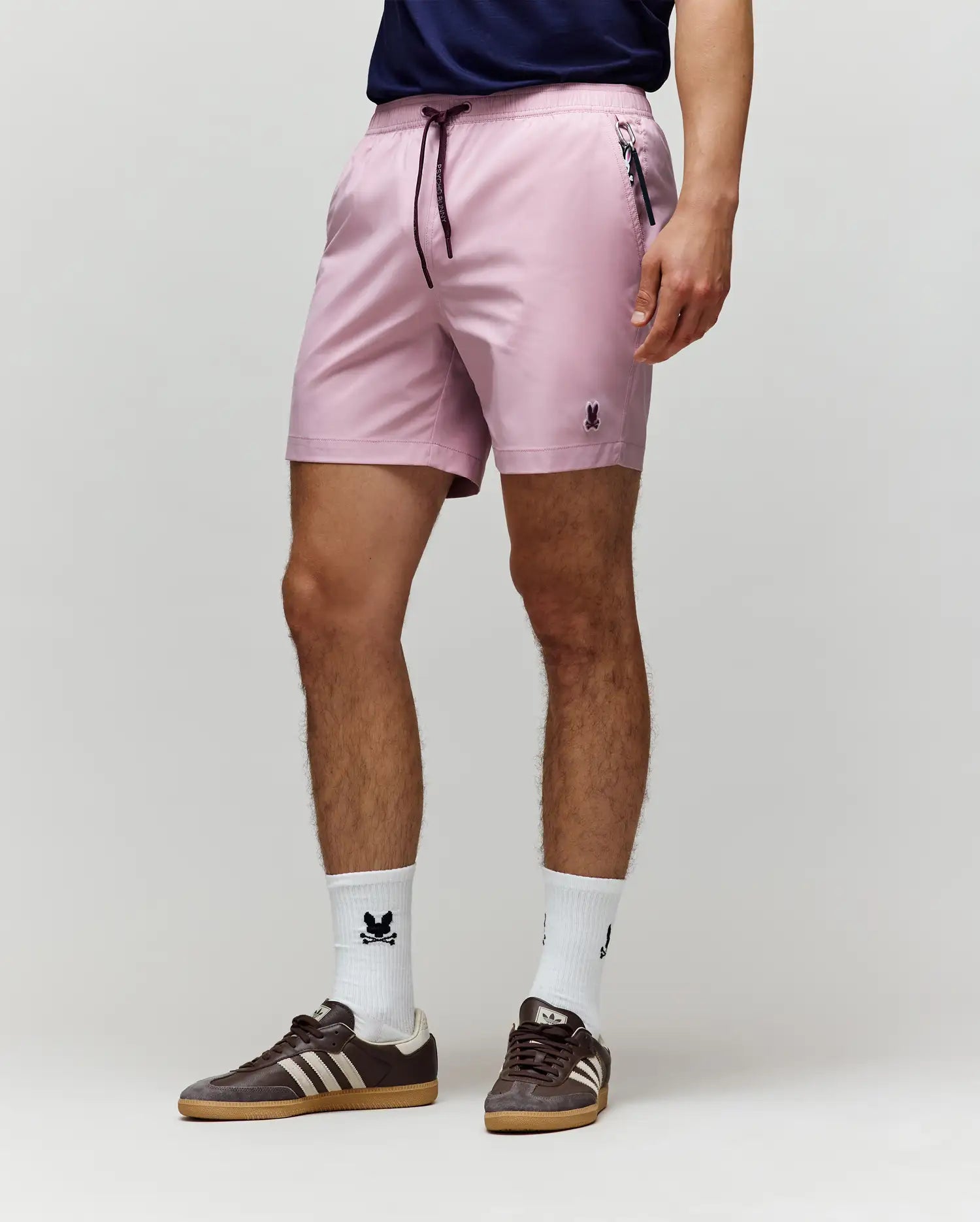 A person wearing Psycho Bunny MENS SAMOS HYDROCHROMIC PRINTED SWIM TRUNK - B6W453F200 in light pink, a navy shirt, white crew socks with a small black logo, and brown Adidas sneakers stands against a plain light gray background.