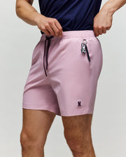 A person wears Psycho Bunny’s MENS SAMOS HYDROCHROMIC PRINTED SWIM TRUNK—light pink shorts with a small bunny logo, bunny keychain, comfy mesh lining—paired with a navy shirt. Face not visible.