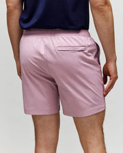 A person is seen from the back wearing Psycho Bunny MENS SAMOS HYDROCHROMIC PRINTED SWIM TRUNK - B6W453F200 in light pink, featuring an elastic waistband, mesh lining, side pocket, and a back pocket with a horizontal opening.