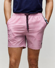 A person stands against a plain light background, hands in pockets, wearing Psycho Bunny MENS SAMOS HYDROCHROMIC PRINTED SWIM TRUNK - B6W453F200 (light pink, quick-dry, drawstring, mesh lining) and a navy blue shirt.
