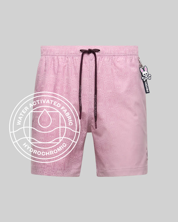 Psycho Bunny MENS SAMOS HYDROCHROMIC PRINTED SWIM TRUNK - B6W453F200: Light pink swim trunks with subtle bunny pattern, mesh lining, dark drawstring, and bunny keychain. Water-activated hydrochromic fabric. White circular graphic overlay. Light gray background.