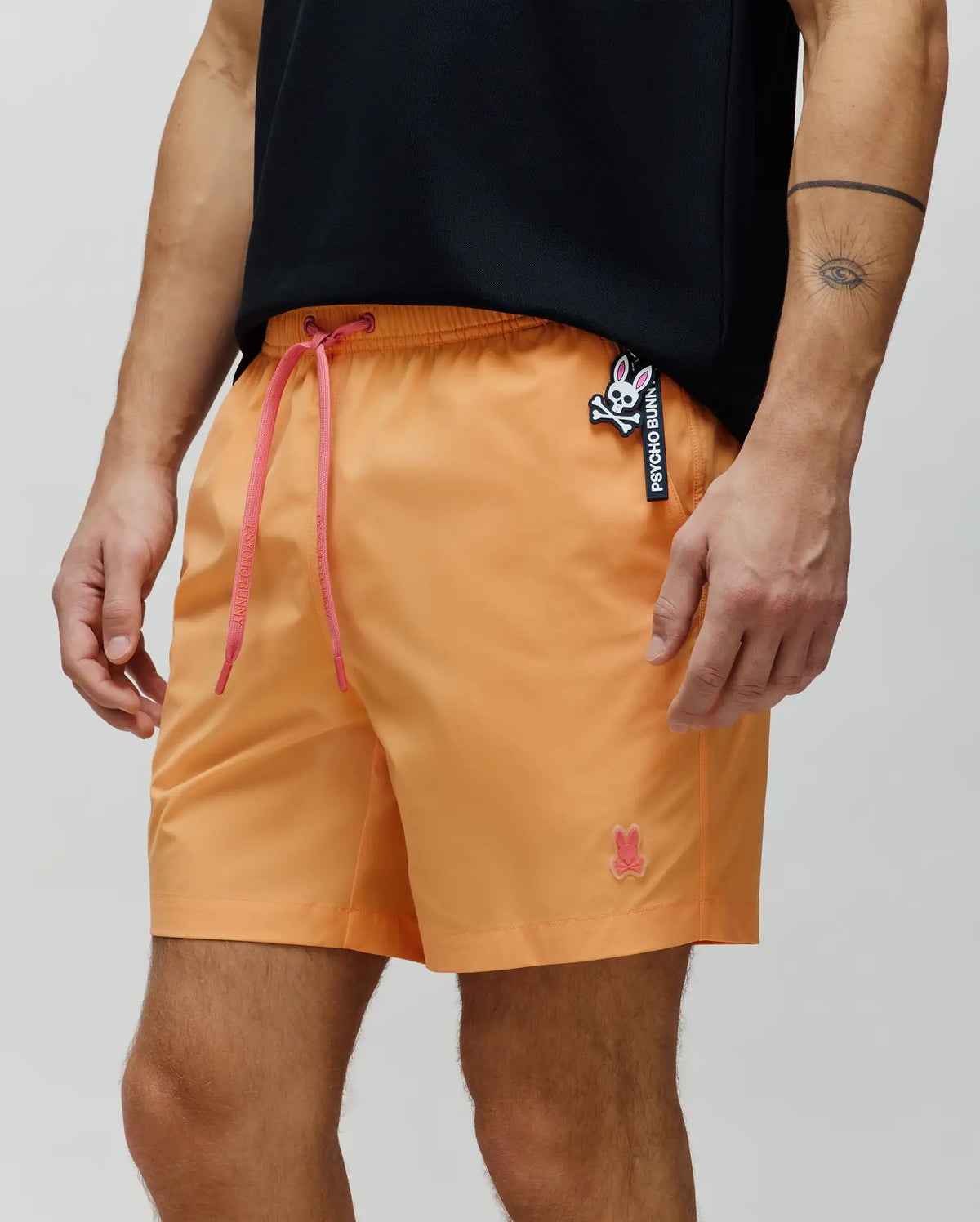 Mens Samos Hydrochromic Printed Swim Trunk in vibrant orange, featuring a relaxed fit, pink drawstrings, a small embroidered pink Psycho Bunny logo on the left leg, and a prominent Psycho Bunny skull tag on the right hip.