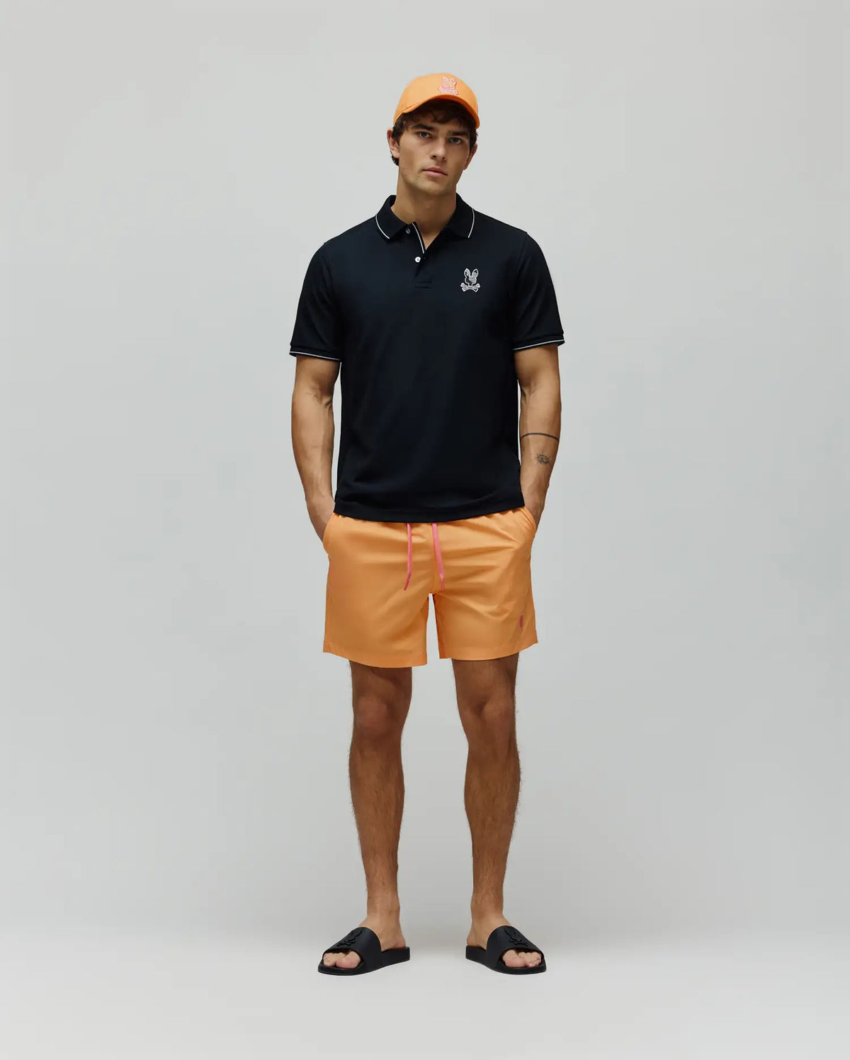 Black pique polo with white tipping and small embroidered Psycho Bunny logo, paired with Mens Samos Hydrochromic Printed Swim Trunk in orange with pink drawstring and small pink logo, an orange cap, and black logo slides.