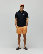 Black pique polo with white tipping and small embroidered Psycho Bunny logo, paired with Mens Samos Hydrochromic Printed Swim Trunk in orange with pink drawstring and small pink logo, an orange cap, and black logo slides.