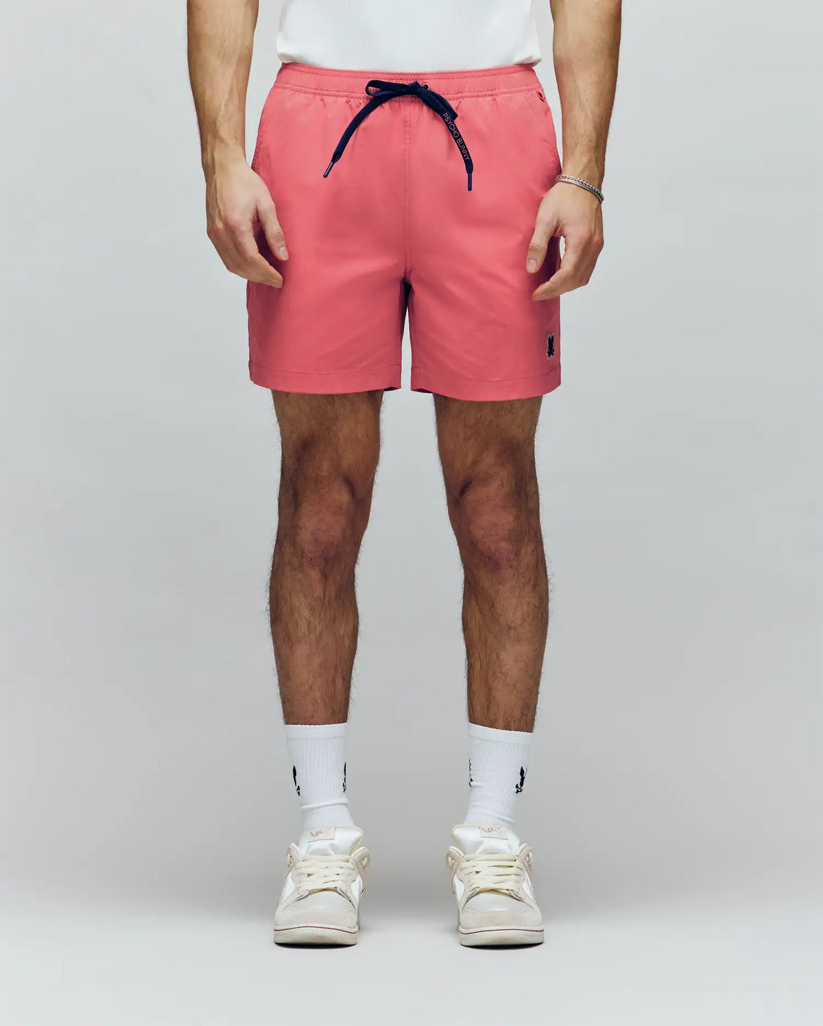 A person models Psycho Bunny's MENS SAMOS HYDROCHROMIC PRINTED SWIM TRUNK (B6W453F200) in coral pink with a navy drawstring, paired with white socks and sneakers. Only the lower torso and legs appear against a plain light background.