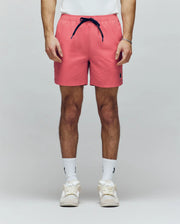 A person models Psycho Bunny's MENS SAMOS HYDROCHROMIC PRINTED SWIM TRUNK (B6W453F200) in coral pink with a navy drawstring, paired with white socks and sneakers. Only the lower torso and legs appear against a plain light background.