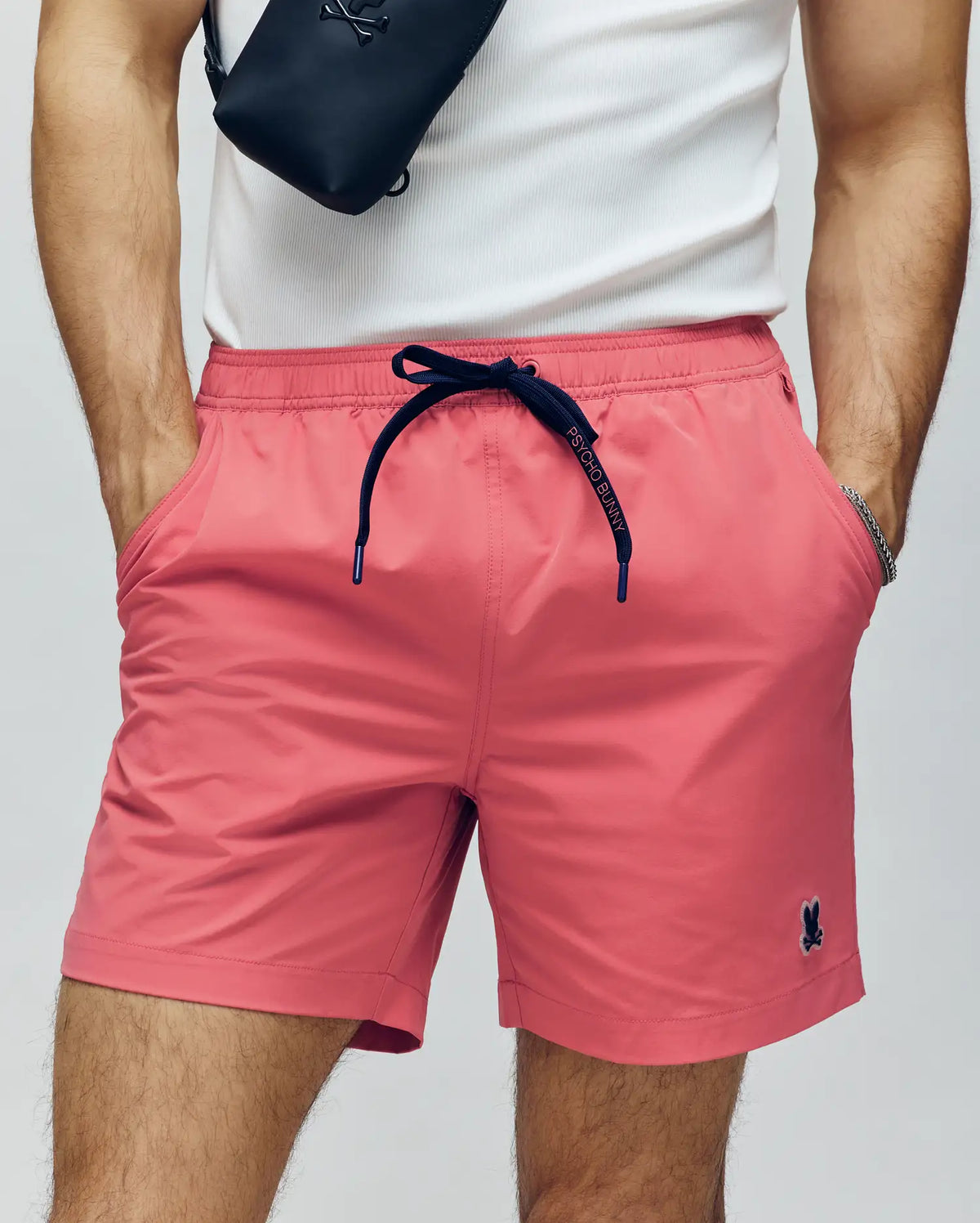 A man wears a white tank top, Psycho Bunny MENS SAMOS HYDROCHROMIC PRINTED SWIM TRUNK shorts with an embroidered logo, a black waterproof crossbody bag, and a silver bracelet. His hands are in his pockets.