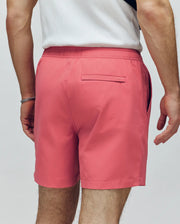 A person, seen from the back, wears Psycho Bunny’s MENS SAMOS HYDROCHROMIC PRINTED SWIM TRUNK (B6W453F200) in salmon pink with a white sleeveless top. The trunks have side and rear pockets and may feature hydrochromic technology.